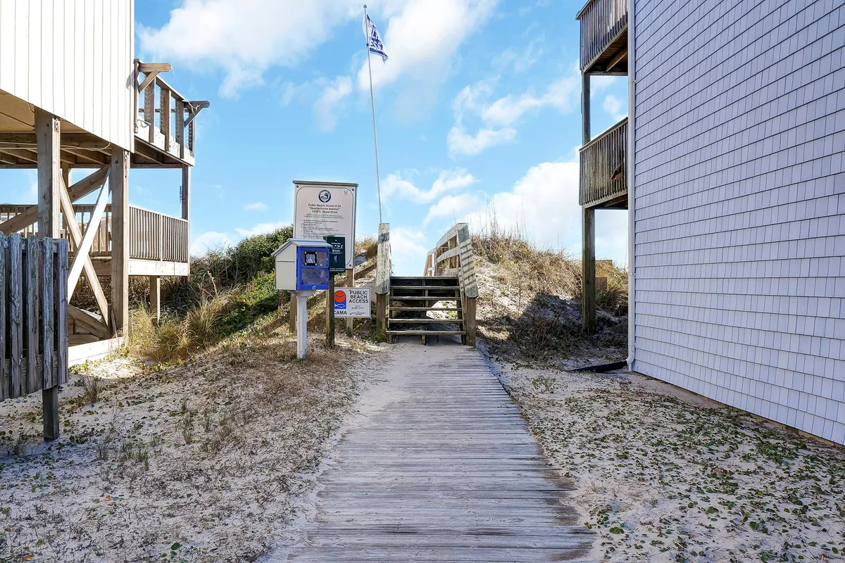 A wooden walkway with stairs leads directly to the beach, flanked by dunes and a flag-topped signpost, symbolizing the easy transition from the property to Surf City's sandy shore. The relaxed atmosphere invites exploration and beachside activities such as yoga or paddleboarding.