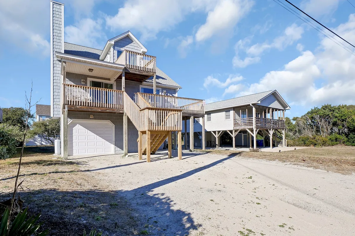 An exterior view of the main house with a welcoming front porch and a large deck, surrounded by greenery and an adjacent building. The scene is set for family gatherings, relaxing with morning coffee, or enjoying sunset views from the deck, emphasizing the comfort and sociability of the space.
