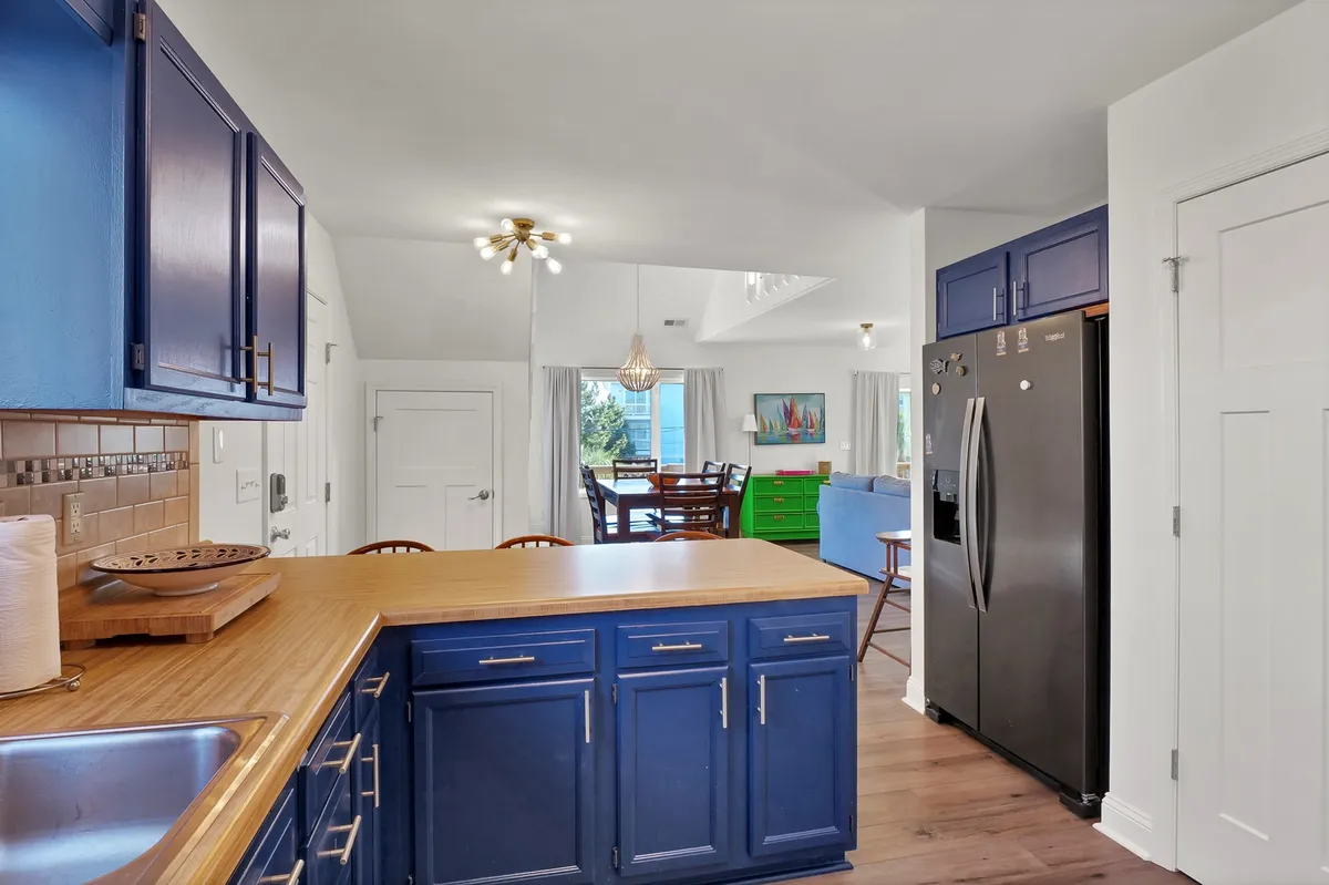 A bright, open-plan space seamlessly connects the kitchen, dining, and living areas. The kitchen features striking blue cabinetry, a warm wooden countertop, and is adjacent to a dining area furnished with colorful chairs. The space is styled with playful coastal touches and plenty of natural light, evoking a lively and welcoming atmosphere.
