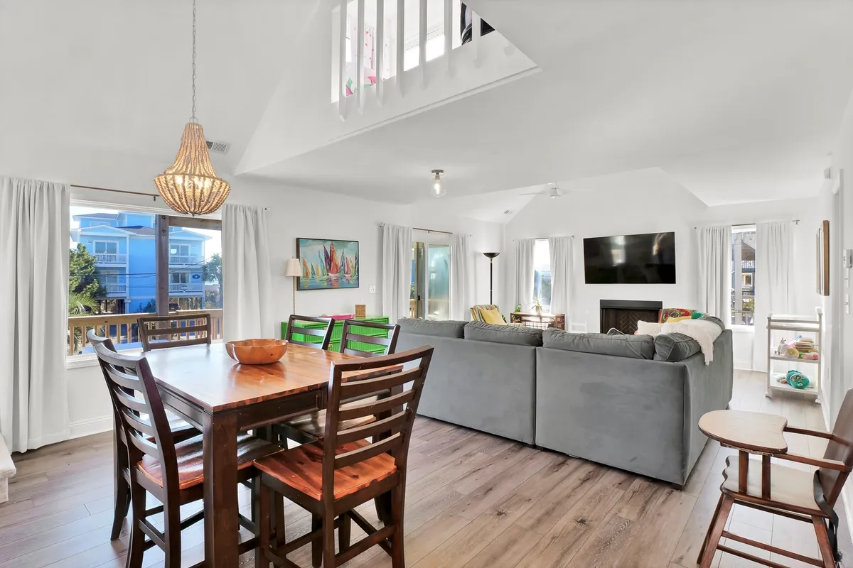 A cozy yet spacious shared area featuring a wooden dining table surrounded by chairs, a comfortable gray sofa, large windows that flood the room with natural light, a stylish pendant light, and a wall-mounted TV. The setup is perfect for shared meals, socializing, or unwinding after a day outdoors.