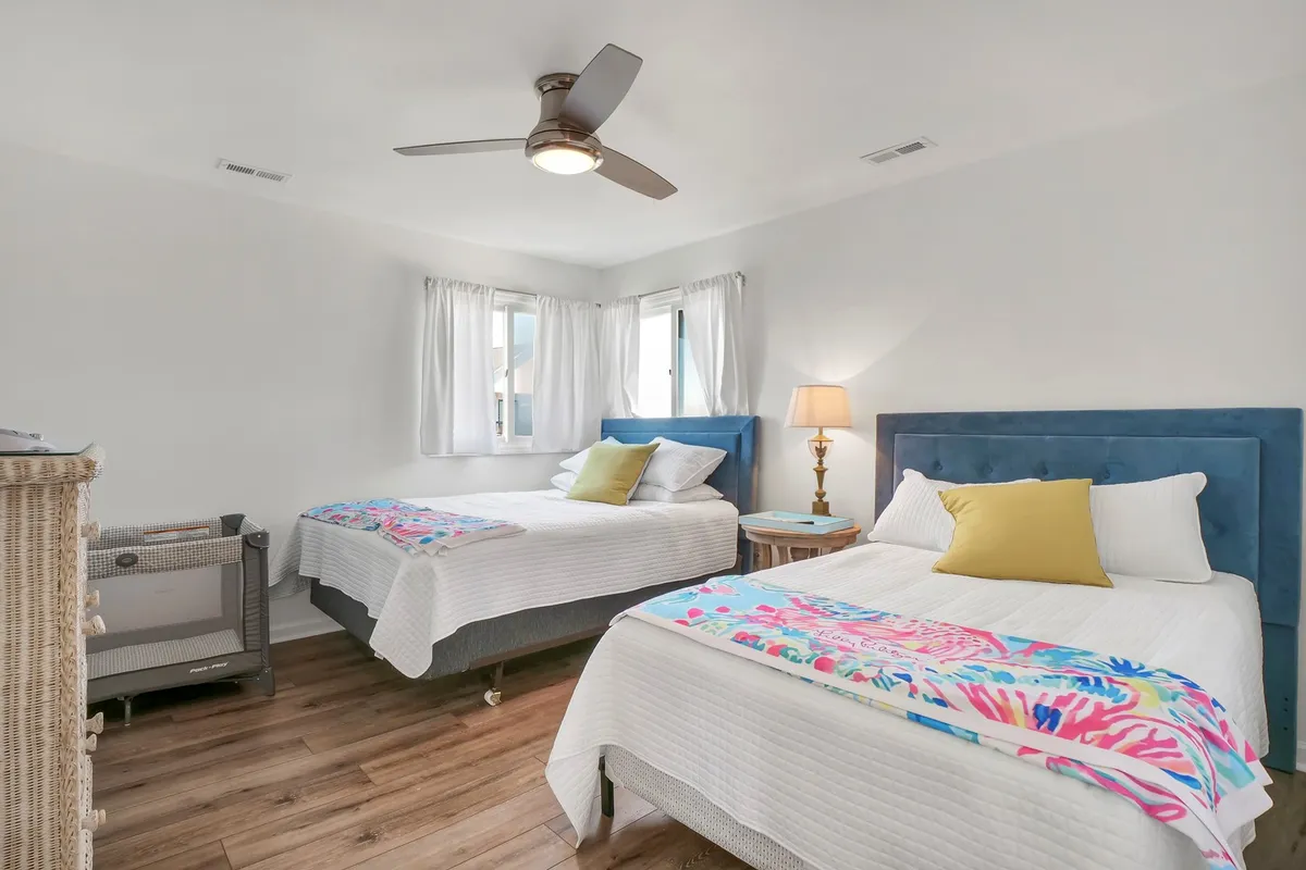 A cozy bedroom featuring two beds with colorful throws, a nightstand with a lamp between them, and a window letting in ample natural light. The layout suggests a room designed for multiple guests or siblings, balancing comfort and shared space.