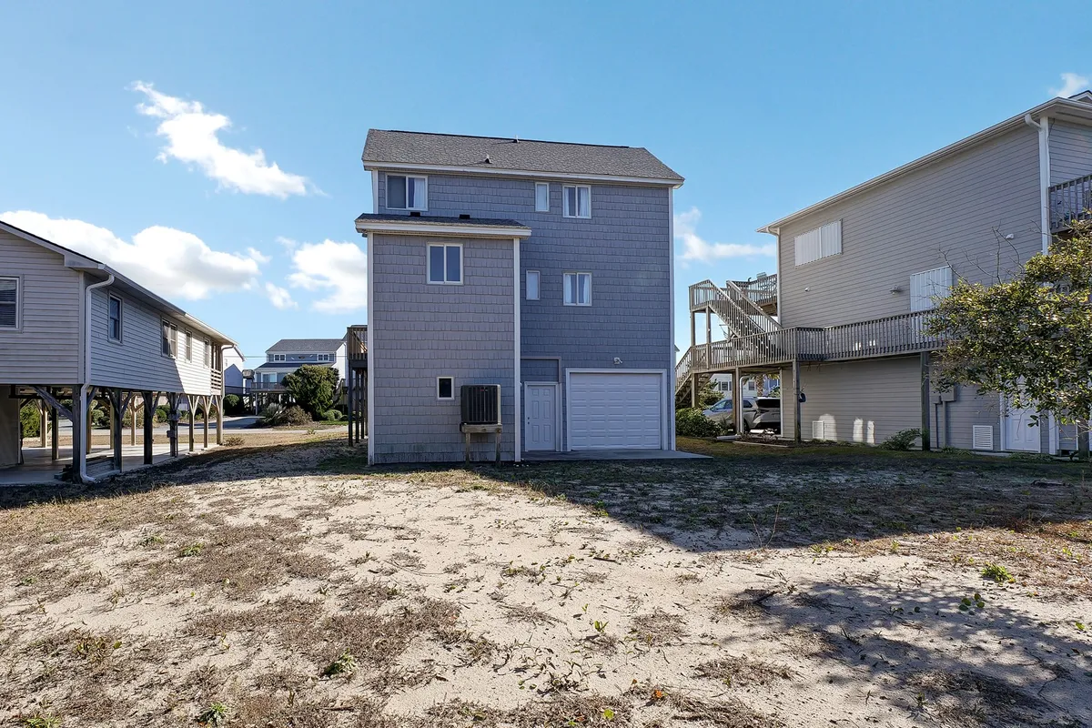 An exterior shot of the two-story house, surrounded by neighboring homes and a spacious sandy area, with a welcoming deck perfect for sunset drinks or group gatherings. The overall scene suggests an environment ideal for outdoor socializing and enjoying evenings together after beach activities.