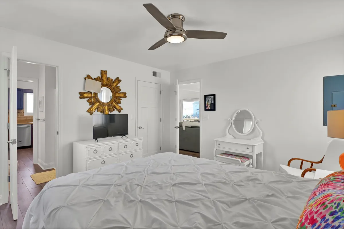 A tranquil, inviting bedroom with a bed dressed in a textured white comforter, a golden sunburst mirror above, a modern ceiling fan, and a small TV atop a dresser. The room feels private yet welcoming, ideal for relaxation or a restful night's sleep.