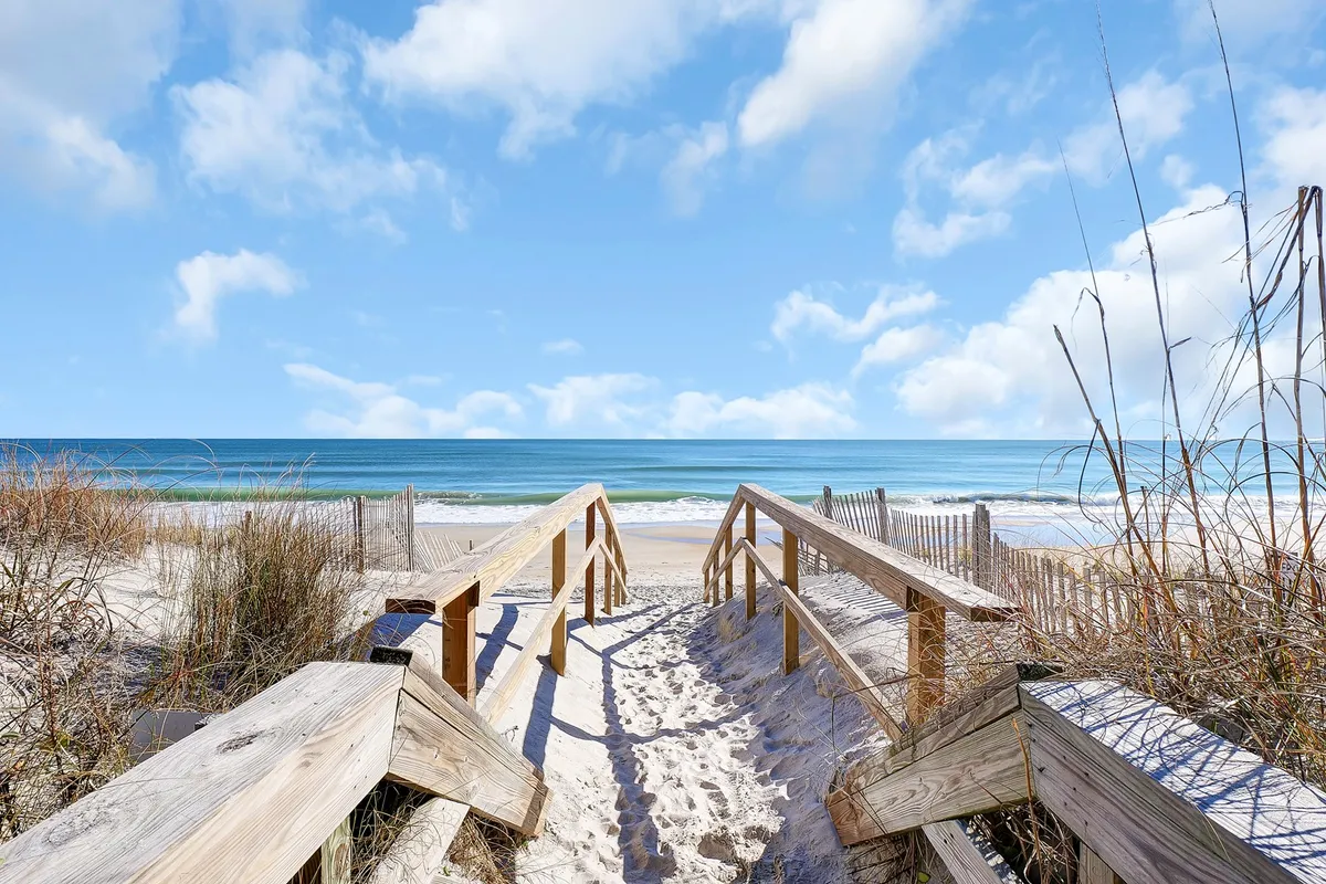 A serene exterior image showing a wooden boardwalk leading through grassy dunes to the ocean beyond. The view includes clear skies, and the beach setting is just steps away, reinforcing the home's immediate access to the coast.