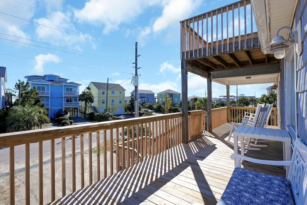 A cheerful exterior shot of a private wooden deck featuring blue chairs and surrounded by colorful neighboring houses. The deck is bathed in sunlight, inviting guests to relax, socialize, or enjoy a meal outdoors. The scene evokes a sense of coastal living and casual comfort.